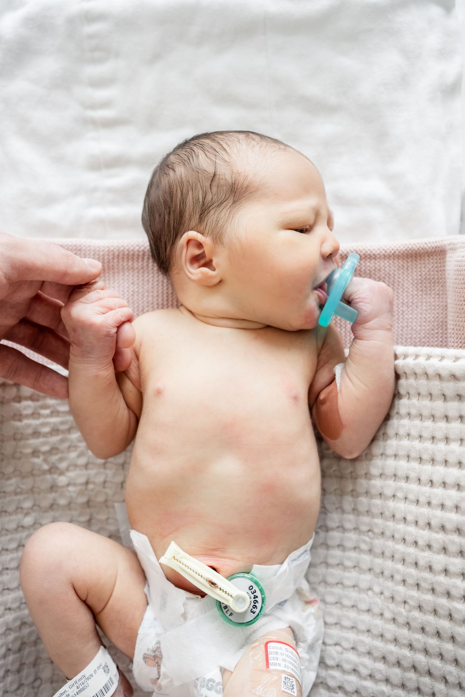 Newborn baby lying on white blanket with blue pacifier and hospital umbilical cord clamp.