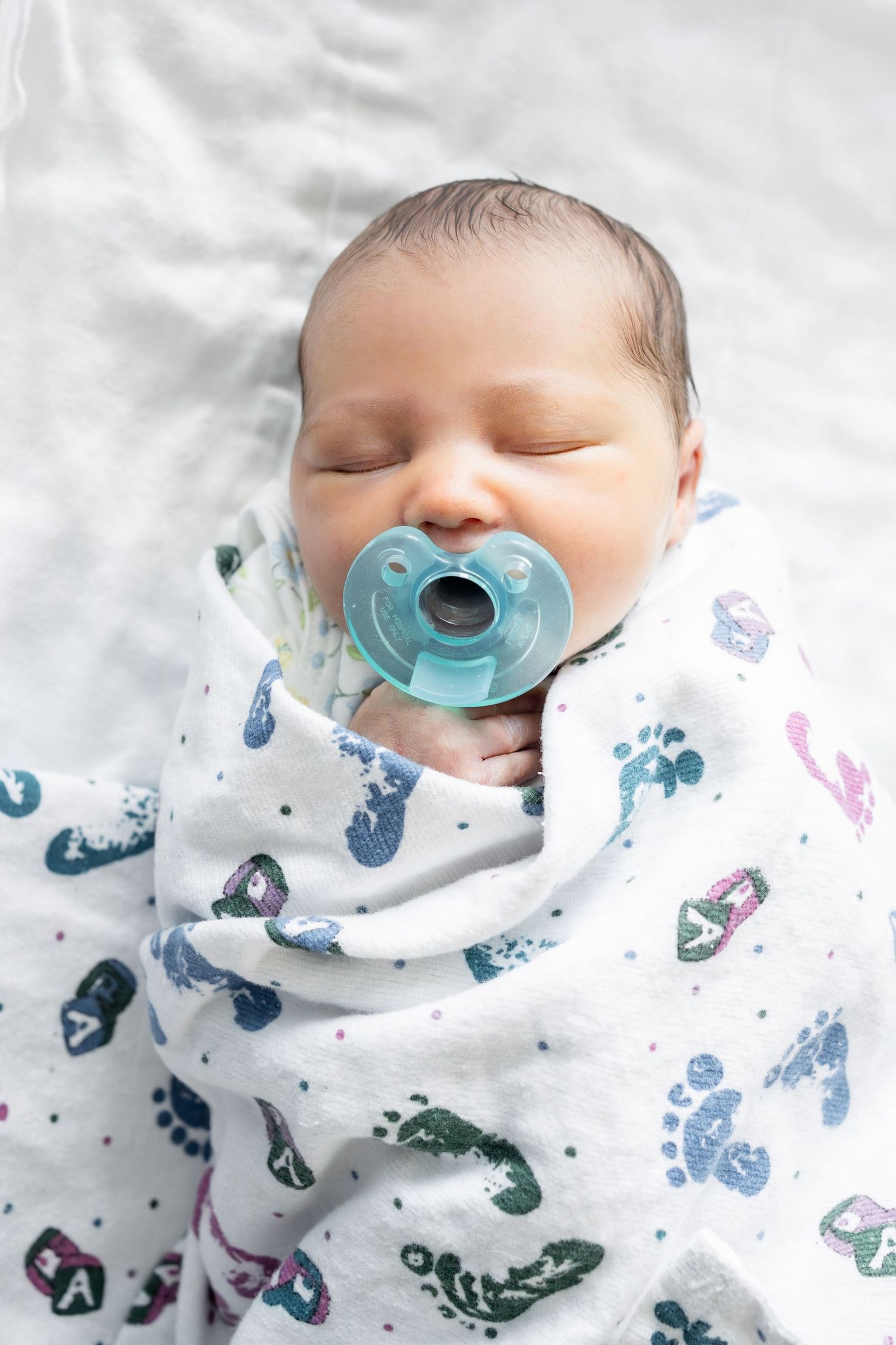Newborn baby sleeping peacefully swaddled in a white blanket with footprint pattern, with a blue pacifier.