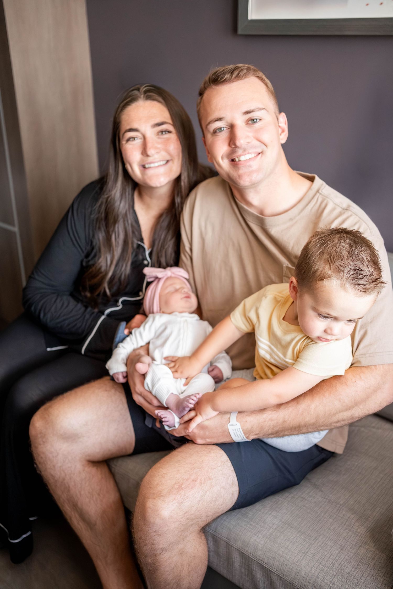 A smiling couple sits together holding a newborn baby and a toddler in a cozy indoor setting.