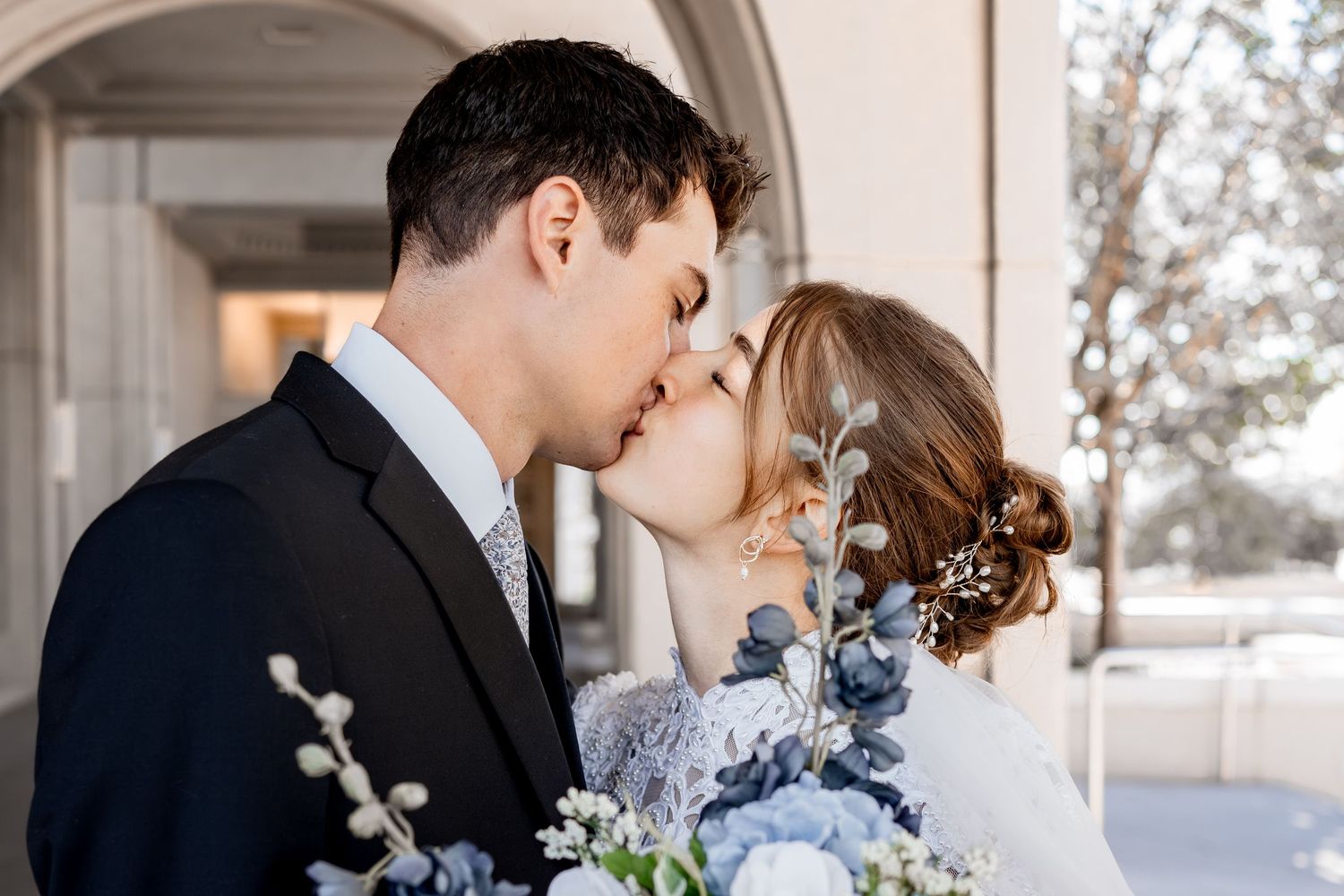 Bride and groom share a romantic kiss under a stone archway, she holds a blue and white floral wedding bouquet and wears a hair accessory.