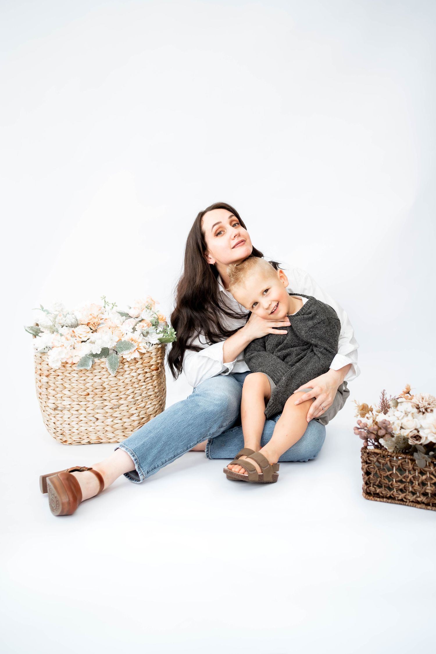 Smiling mother with long dark hair sits in jeans hugging her young son, with woven baskets of dried flowers on a white studio background.