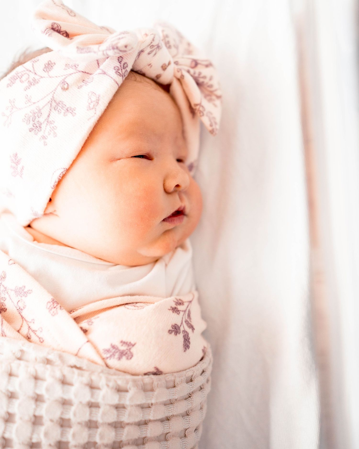 Newborn baby swaddled in pink floral wrap with matching bow headband, sleeping peacefully on white blanket.