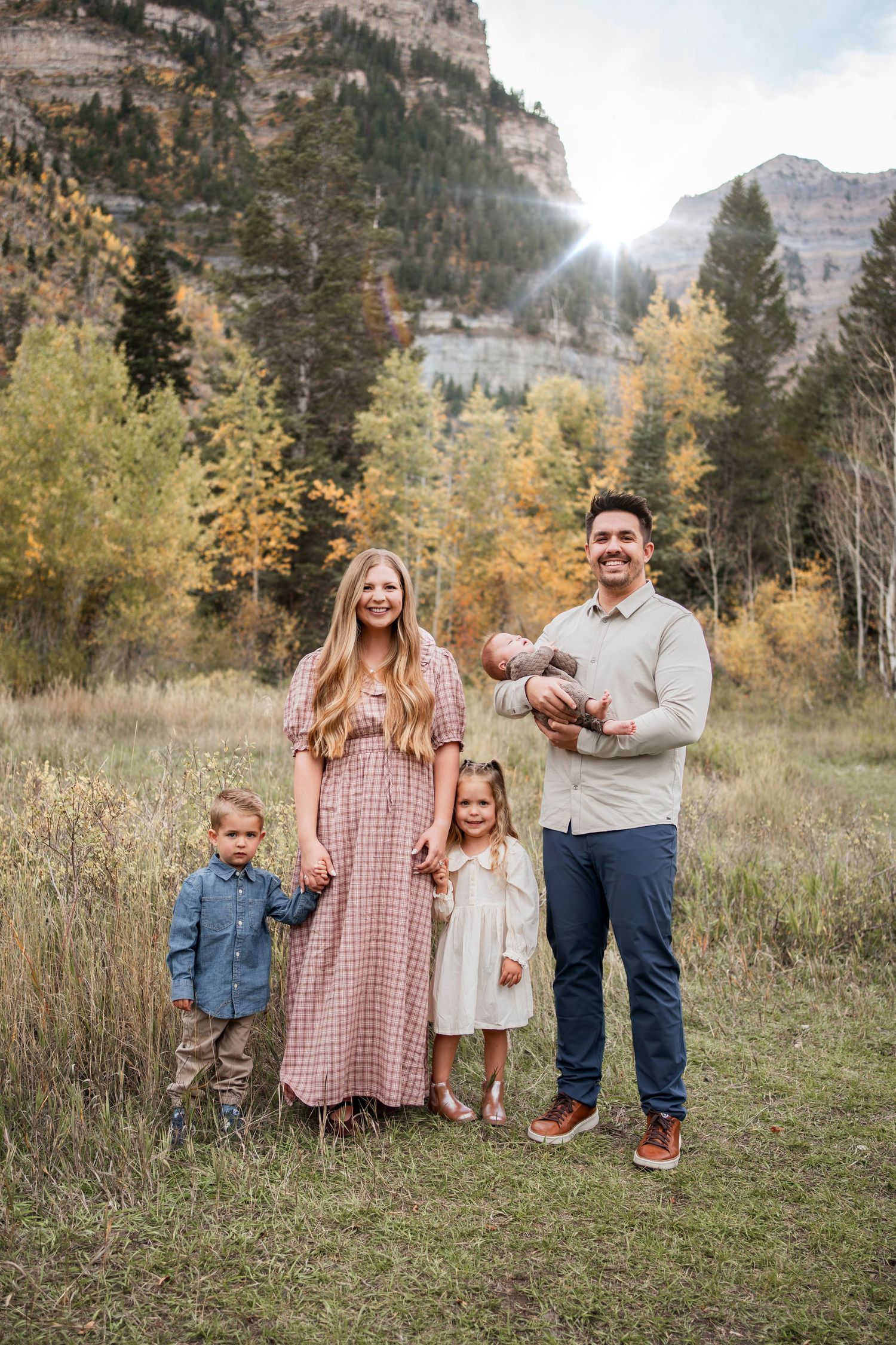 Family of five posing in a golden autumn meadow with a mountain backdrop, parents with three young children, and a baby held by the father.