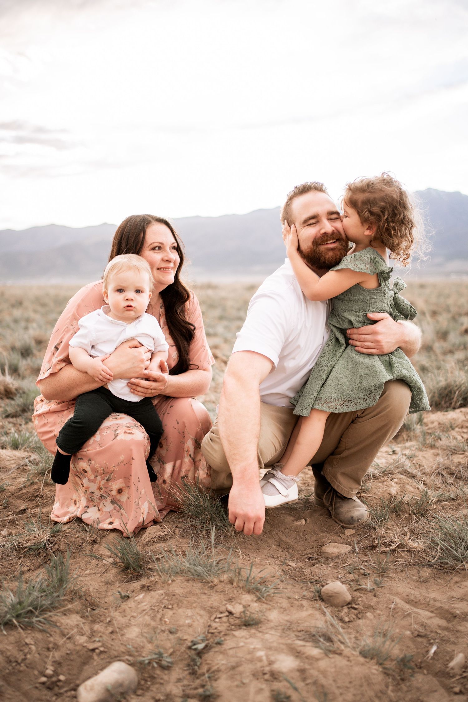 Family of four posing outdoors in desert landscape, mother holding baby, father being hugged by toddler girl.