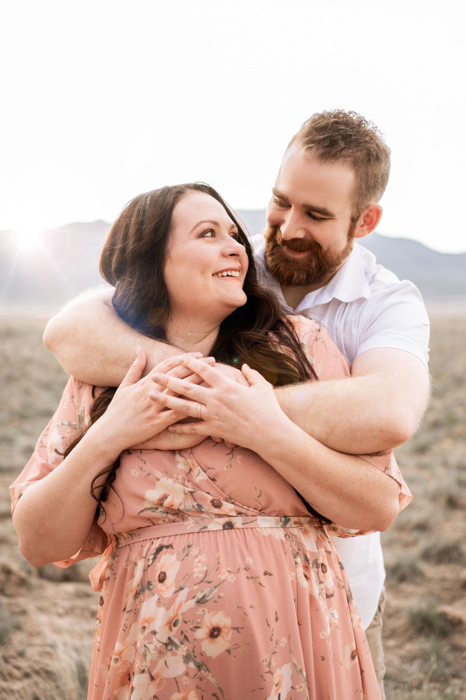 A bearded man embraces a smiling woman in a pink floral dress outdoors, sharing a tender romantic moment.