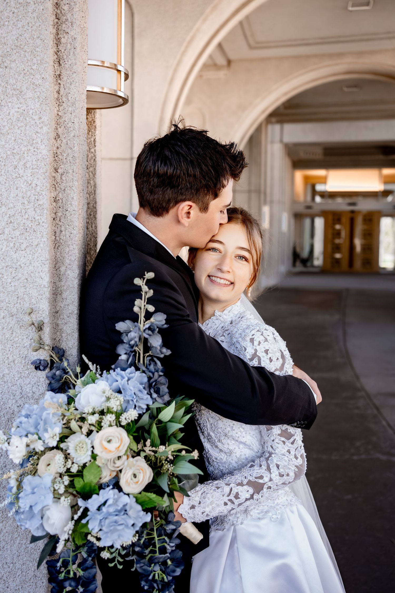 Smiling bride in a lace wedding gown embraces her groom as he kisses her forehead, holding a blue and white floral bouquet.