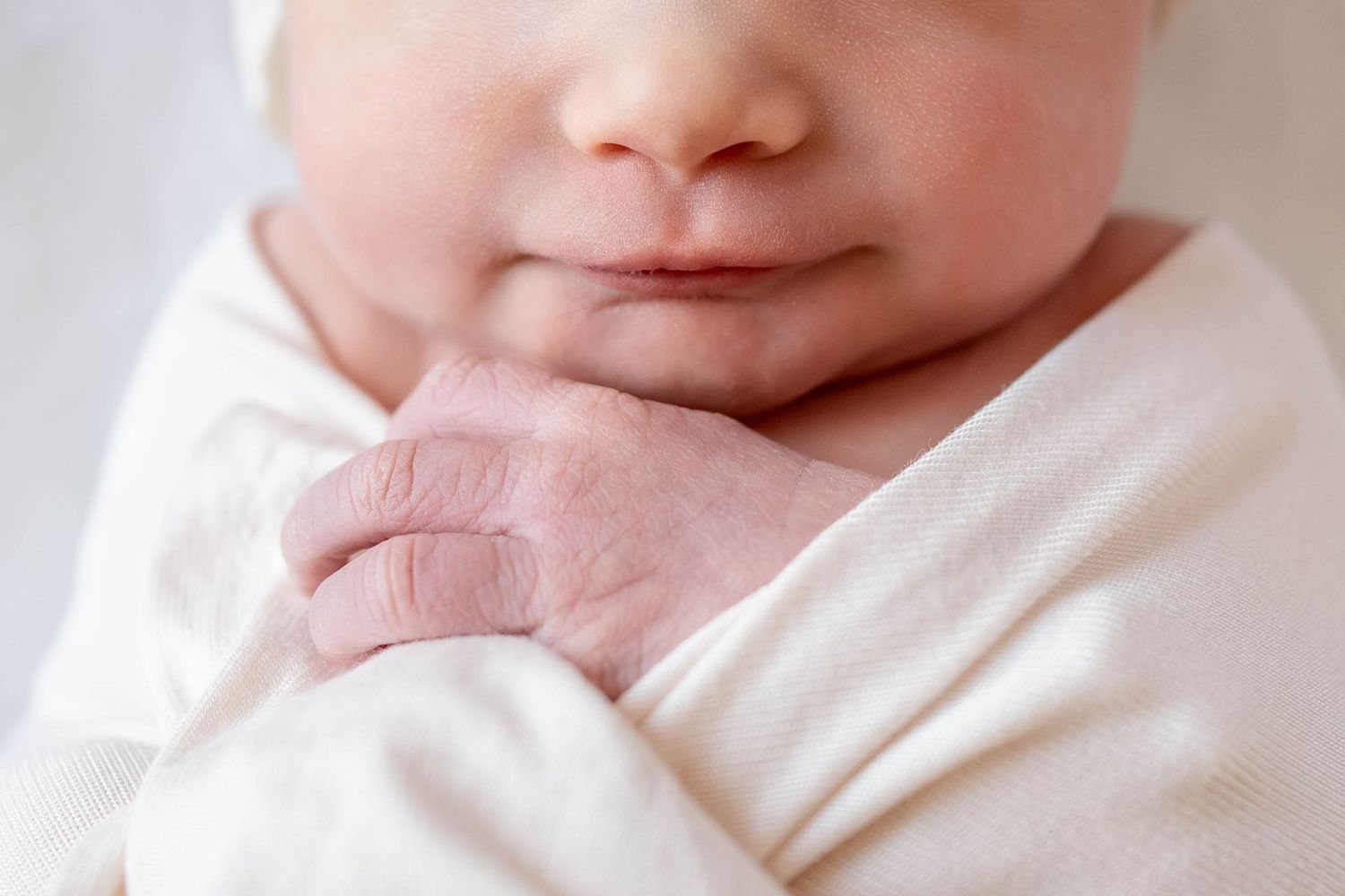 Close-up of a newborn baby's face and tiny hand tucked under the chin, wrapped in a soft cream swaddle blanket.