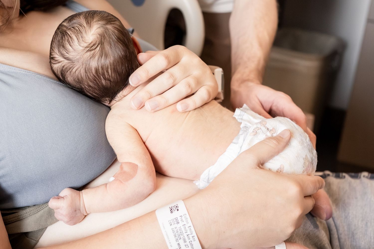 Newborn baby in diaper held against mother's chest for skin-to-skin contact in hospital room.