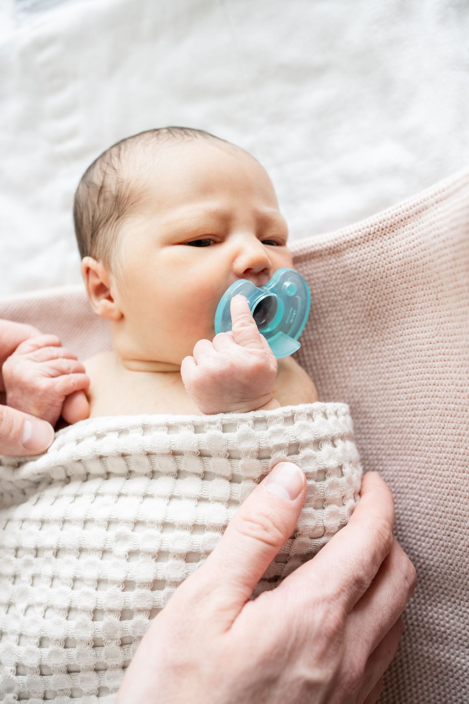 Newborn baby with blue pacifier wrapped in white knit blanket, cradled by adult hands on soft bedding.