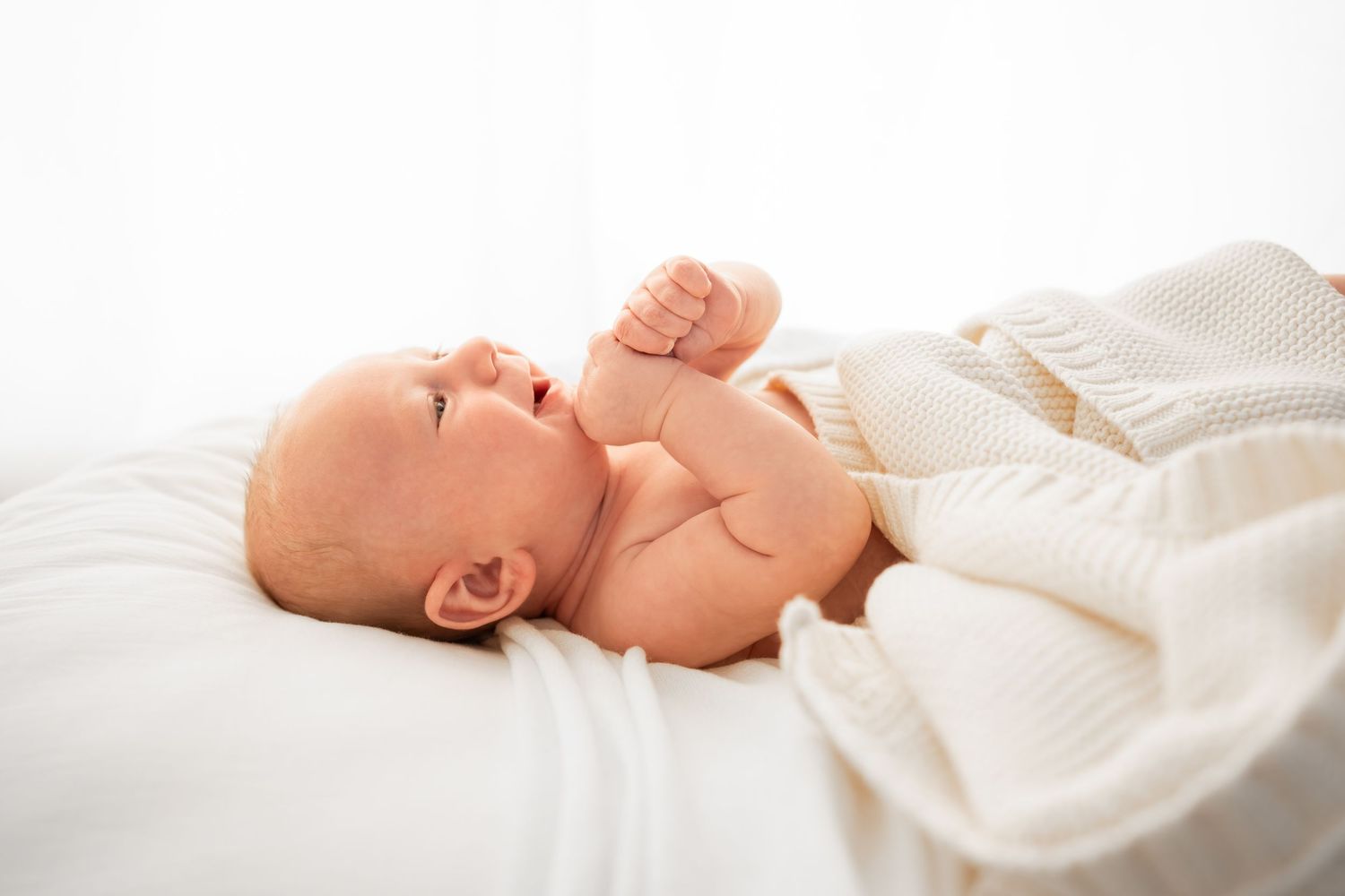 Newborn baby lying on a white cushion with a fist raised near the face, wrapped in a soft cream knit blanket in bright natural light.