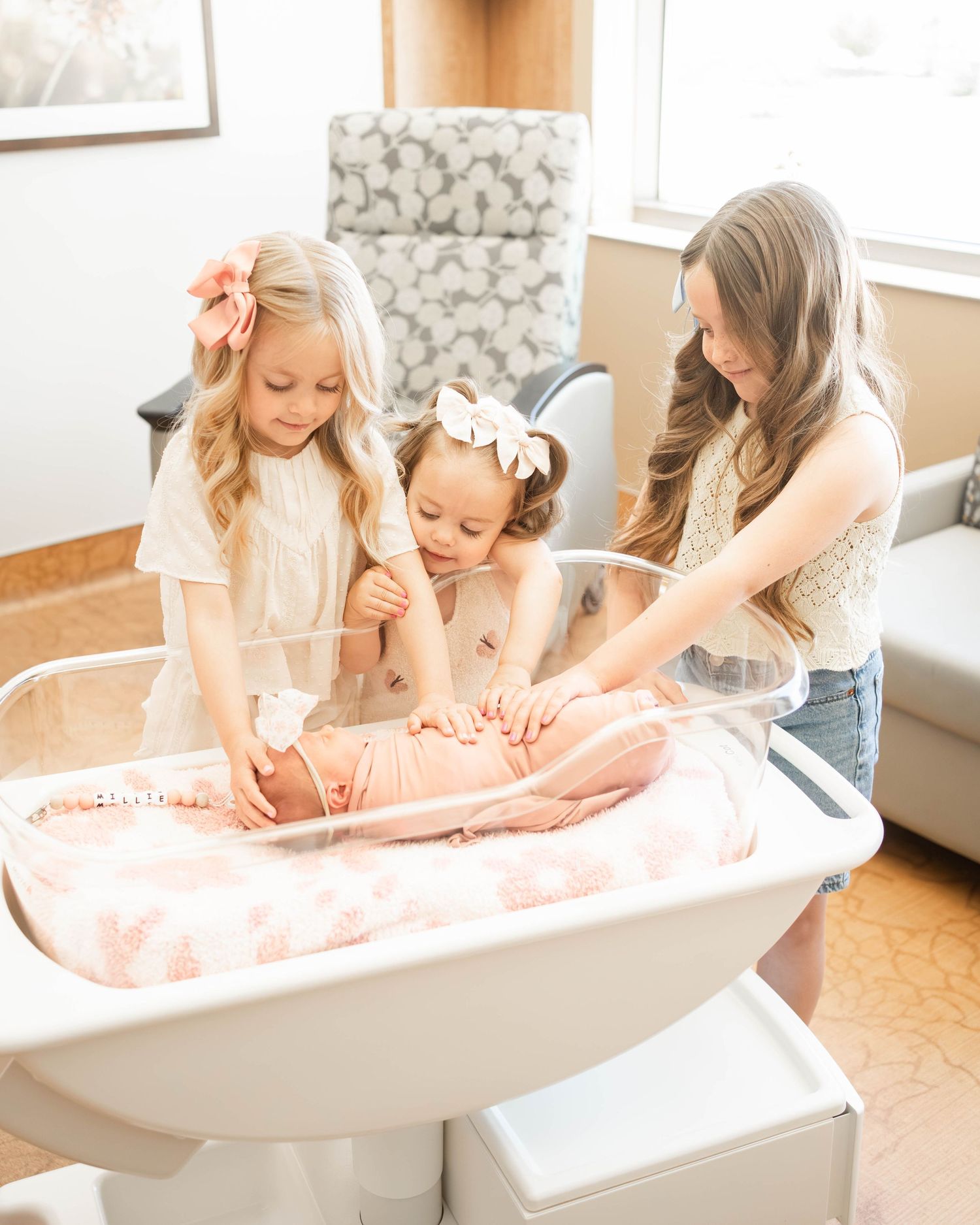 A woman and two young girls in a nursery look at a newborn baby in a clear hospital bassinet.