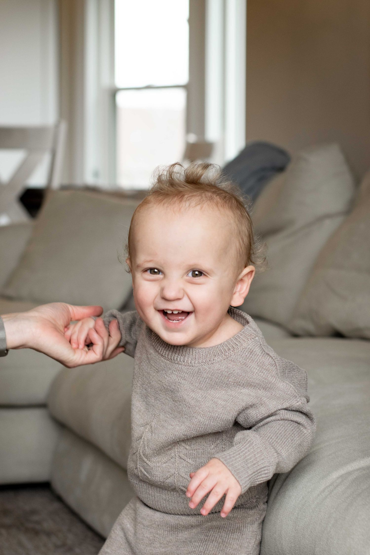Baby in grey sweater laughing joyfully on white bedding.
