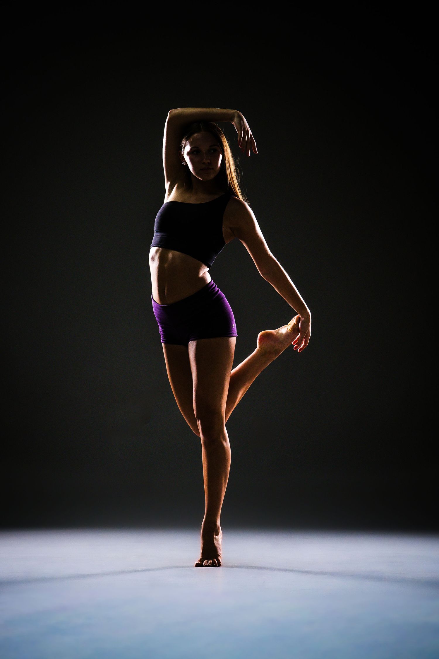A fit woman in purple shorts and black crop top poses gracefully on one leg against a dark background.