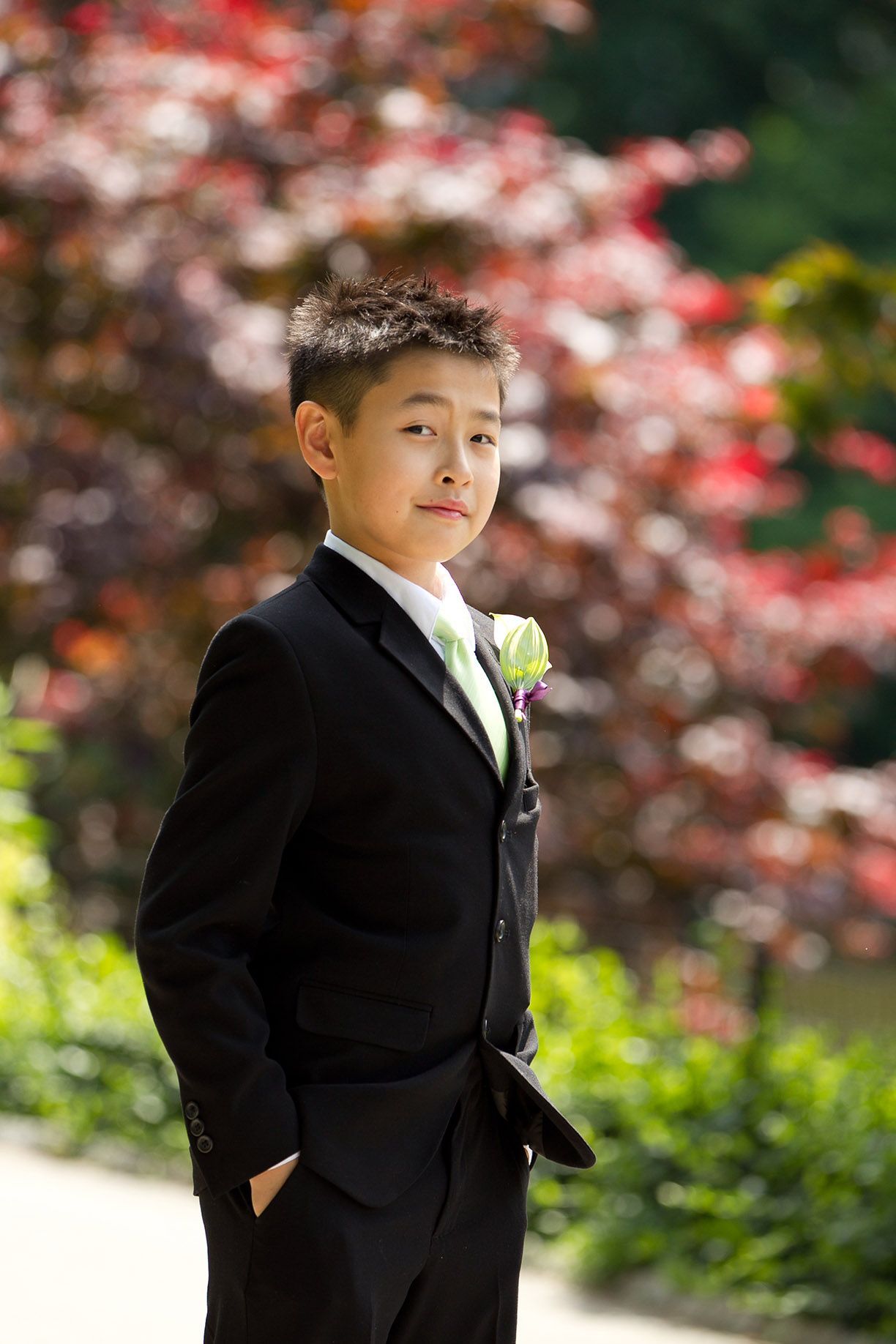 Young person in formal black suit posing in garden with pink flowers.
