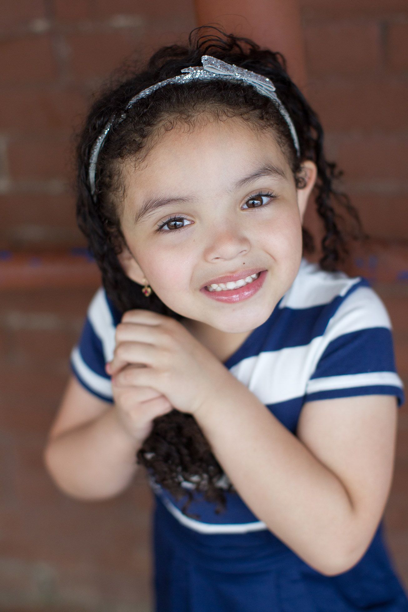 Young child in navy striped dress with sparkly headband against brick wall.