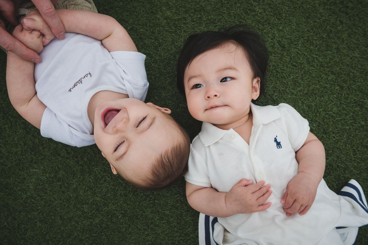 Two babies in white outfits lying on artificial grass surface.