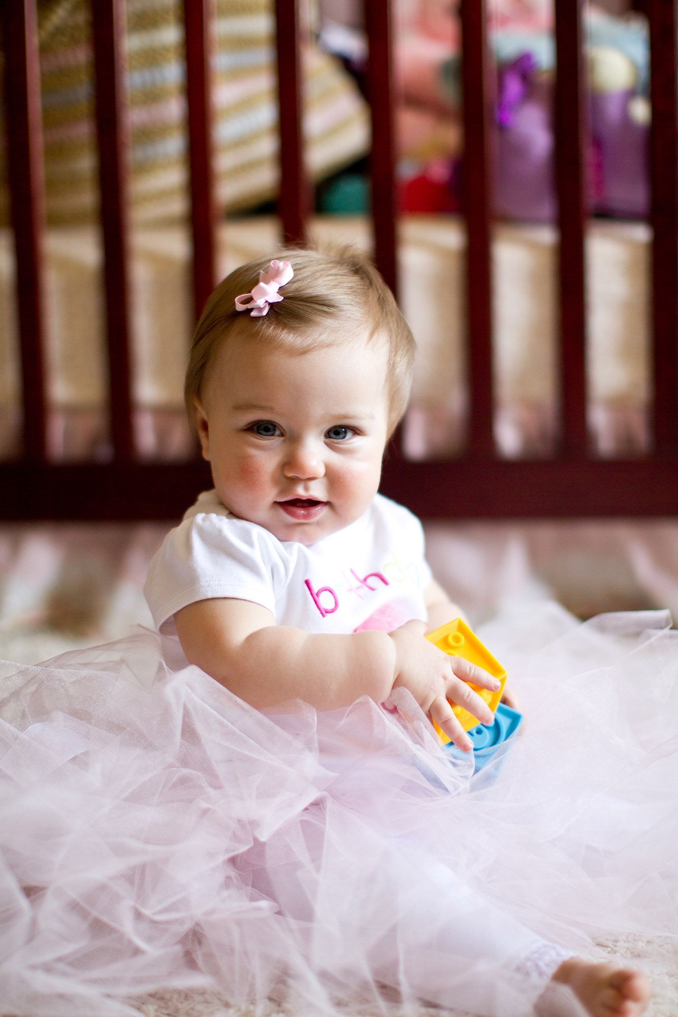 Baby wearing white tulle dress plays with colorful toys in crib.