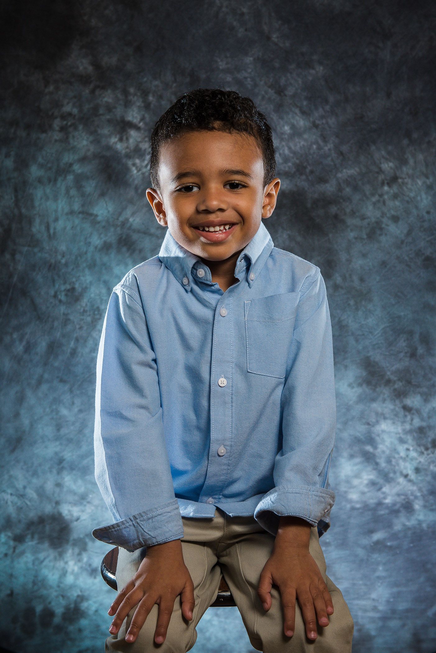 Child in light blue button-down shirt poses on stool against textured gray background.