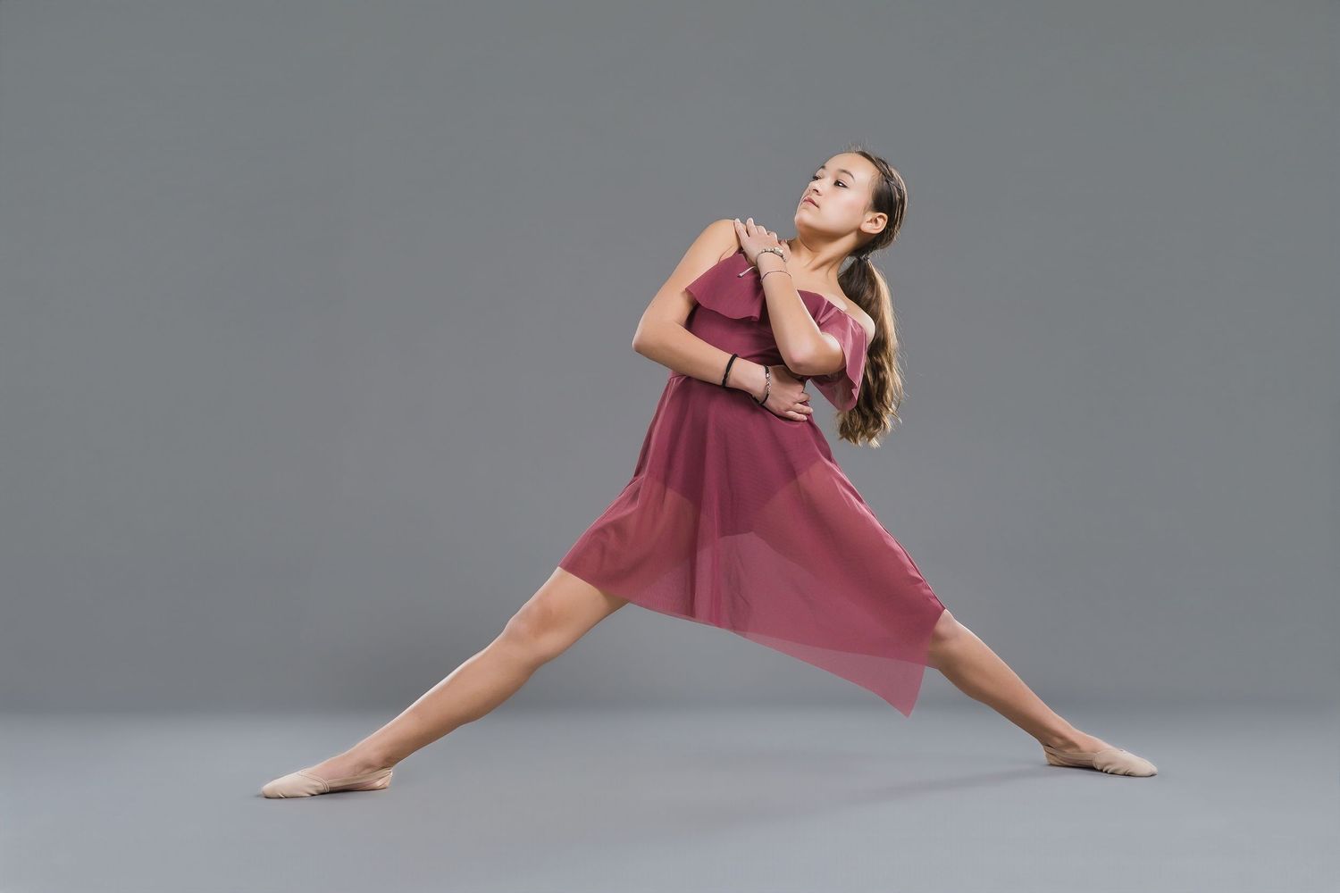 Young female dancer in pink dress performs elegant lunge pose against gray studio background.