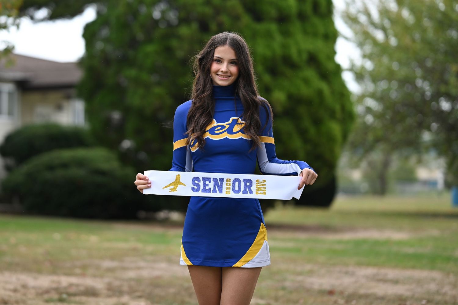 A cheerleader in blue and gold uniform poses outdoors holding a 'SENIORS' sign.
