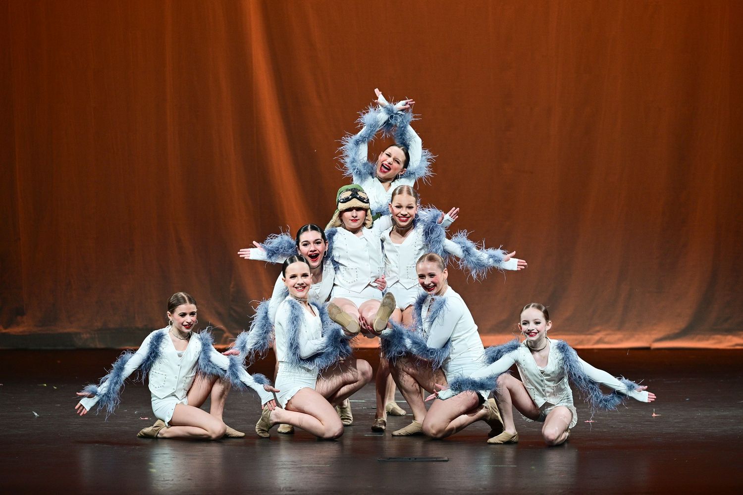 Dance performers in light blue costumes pose in a pyramid formation during a stage performance.