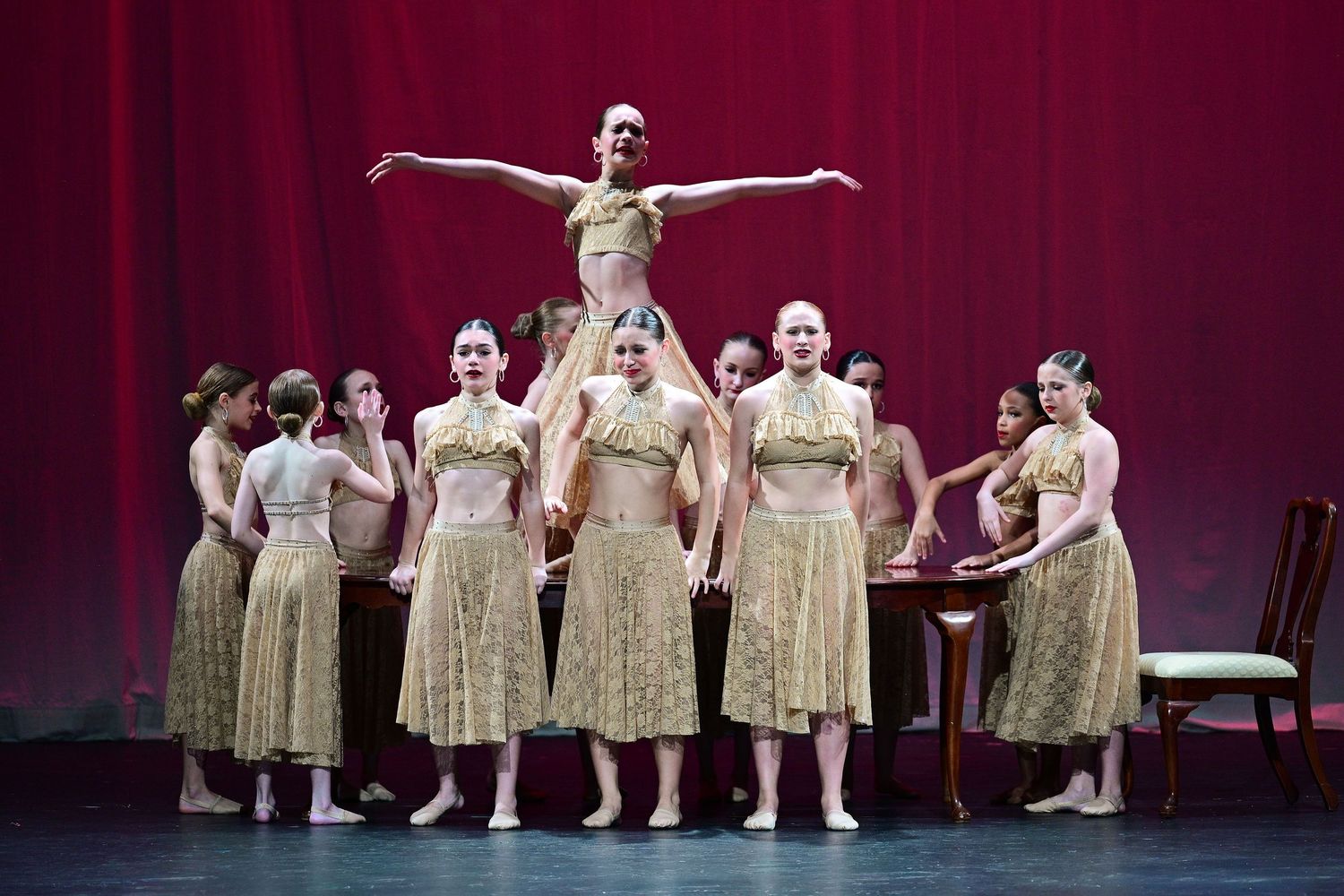 A ballet ensemble in gold dresses performs on stage with a dancer lifted overhead before a red curtain.