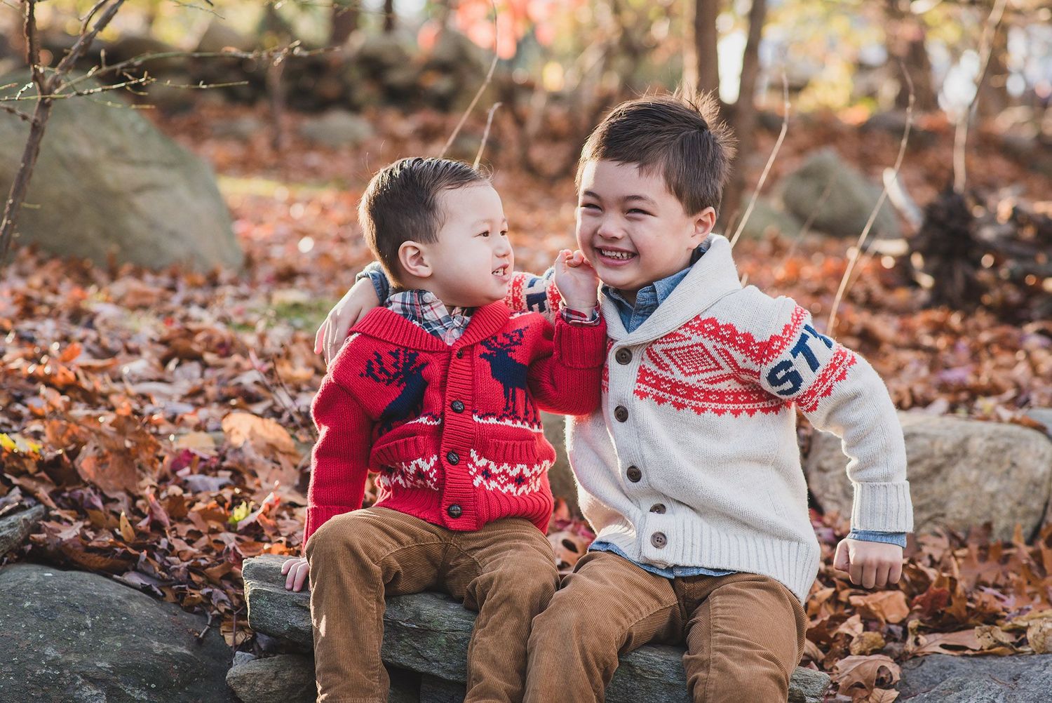 Two young children play together in autumn leaves wearing cozy sweaters and winter accessories.