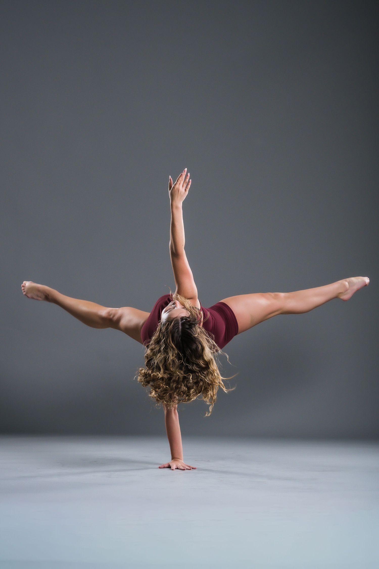 Flexible female dancer performing a one-handed handstand split in a red outfit against a gray background.