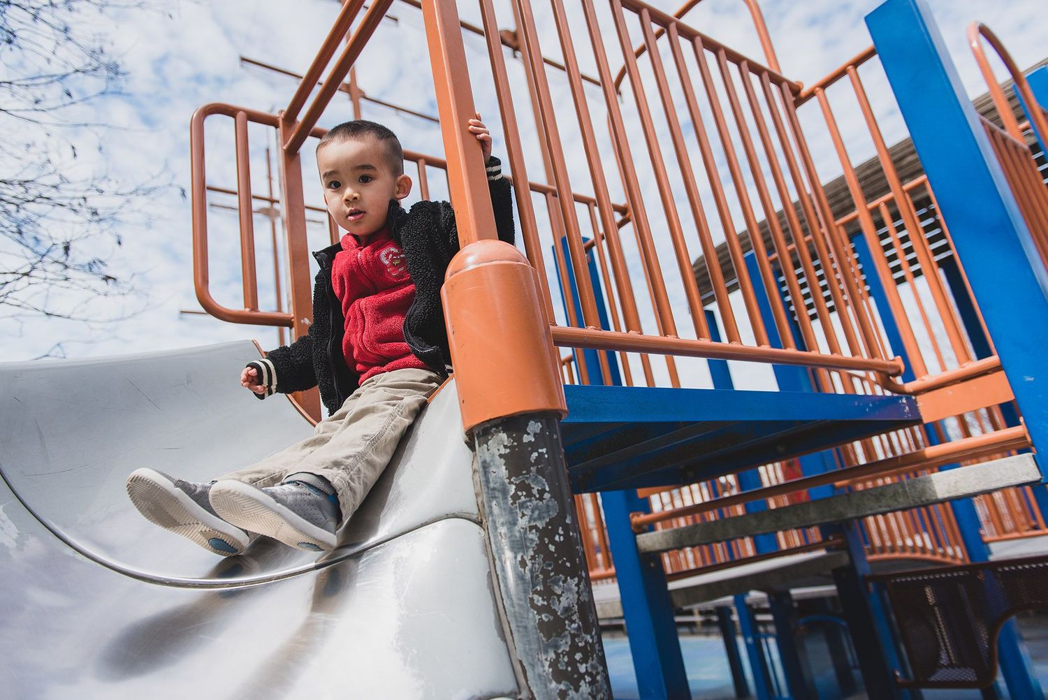 Child playing on orange and blue playground equipment with metal slide on sunny day.