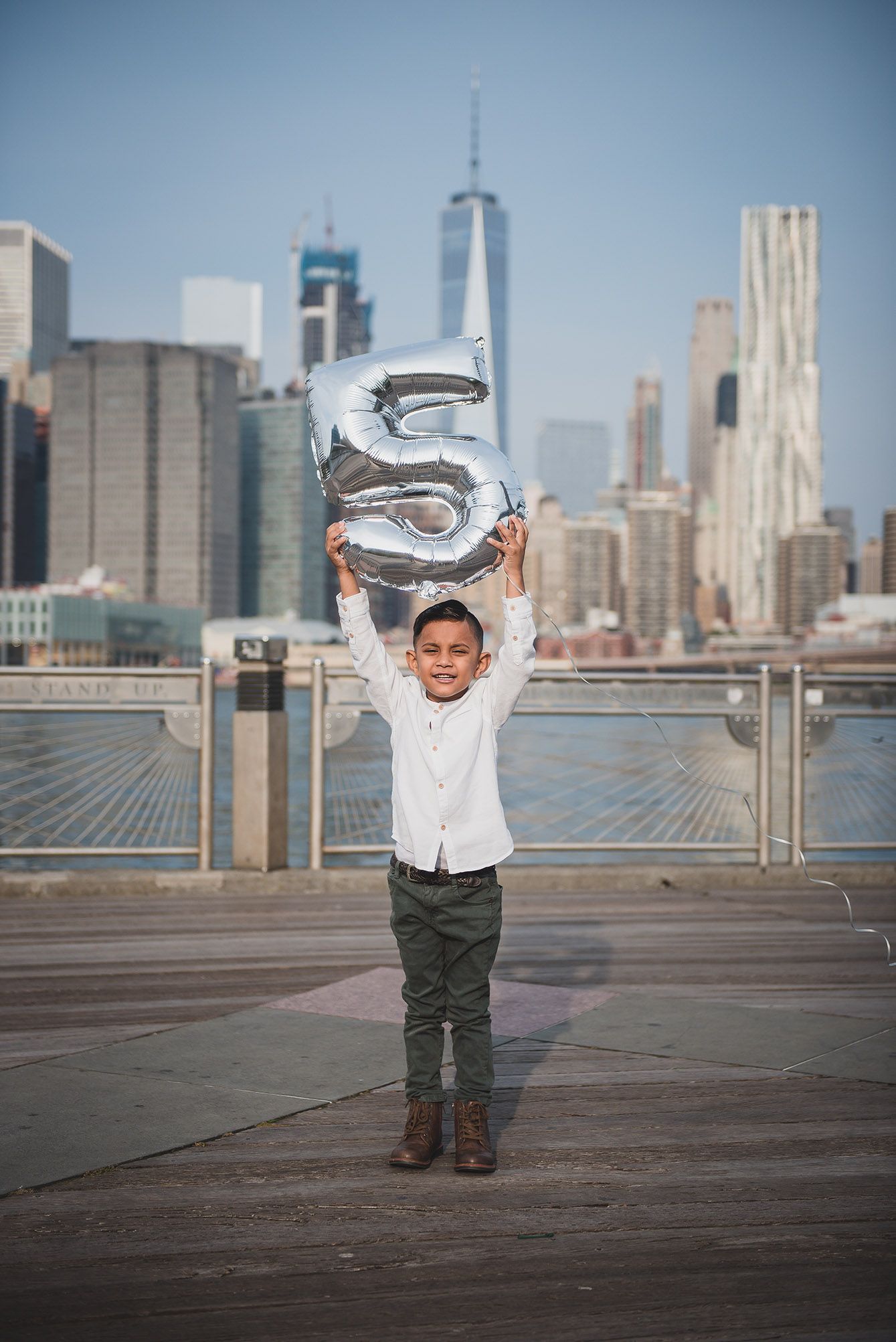 Small child in white shirt and green pants poses against NYC skyline on wooden deck.