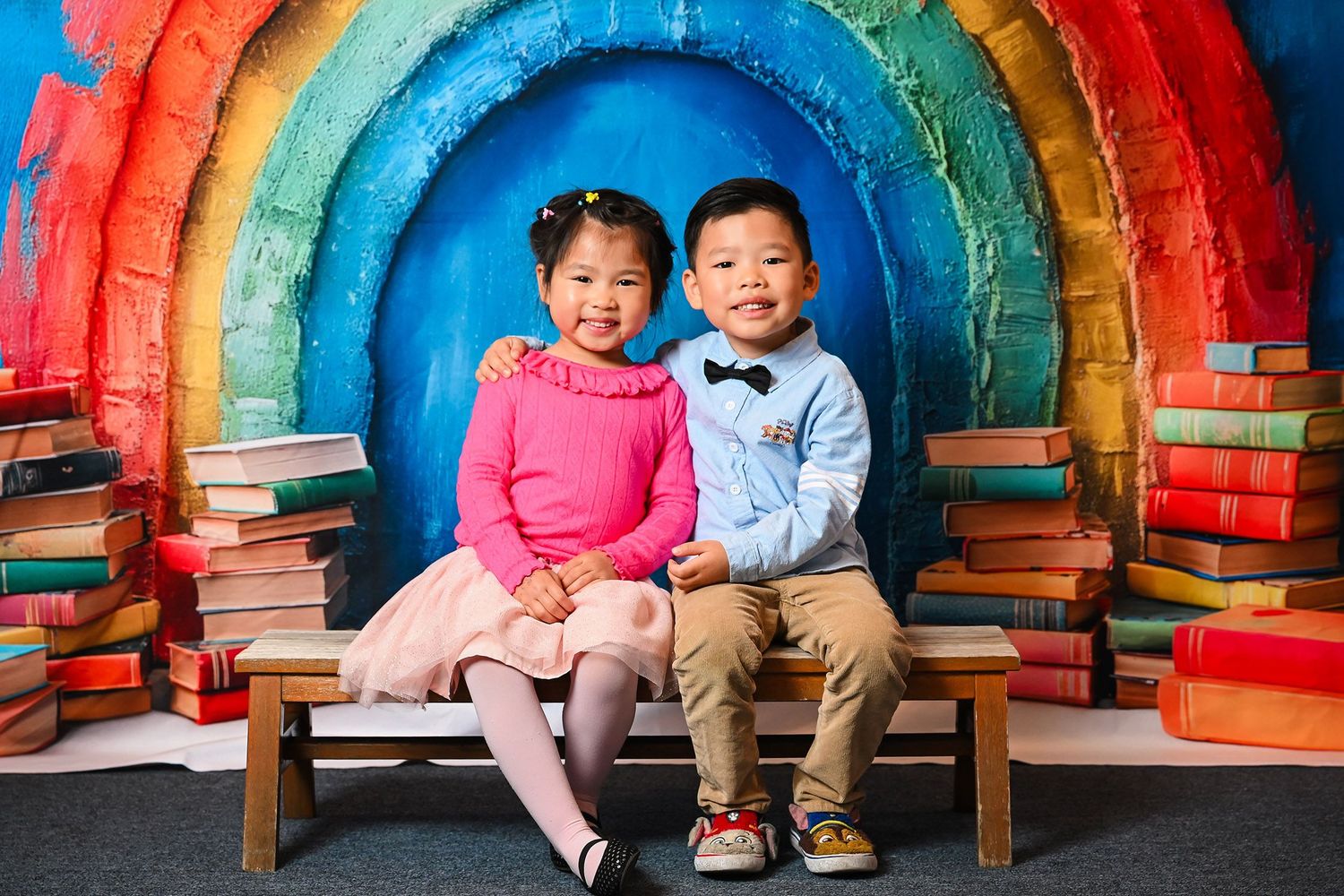 Children sit together on wooden bench against colorful rainbow backdrop.