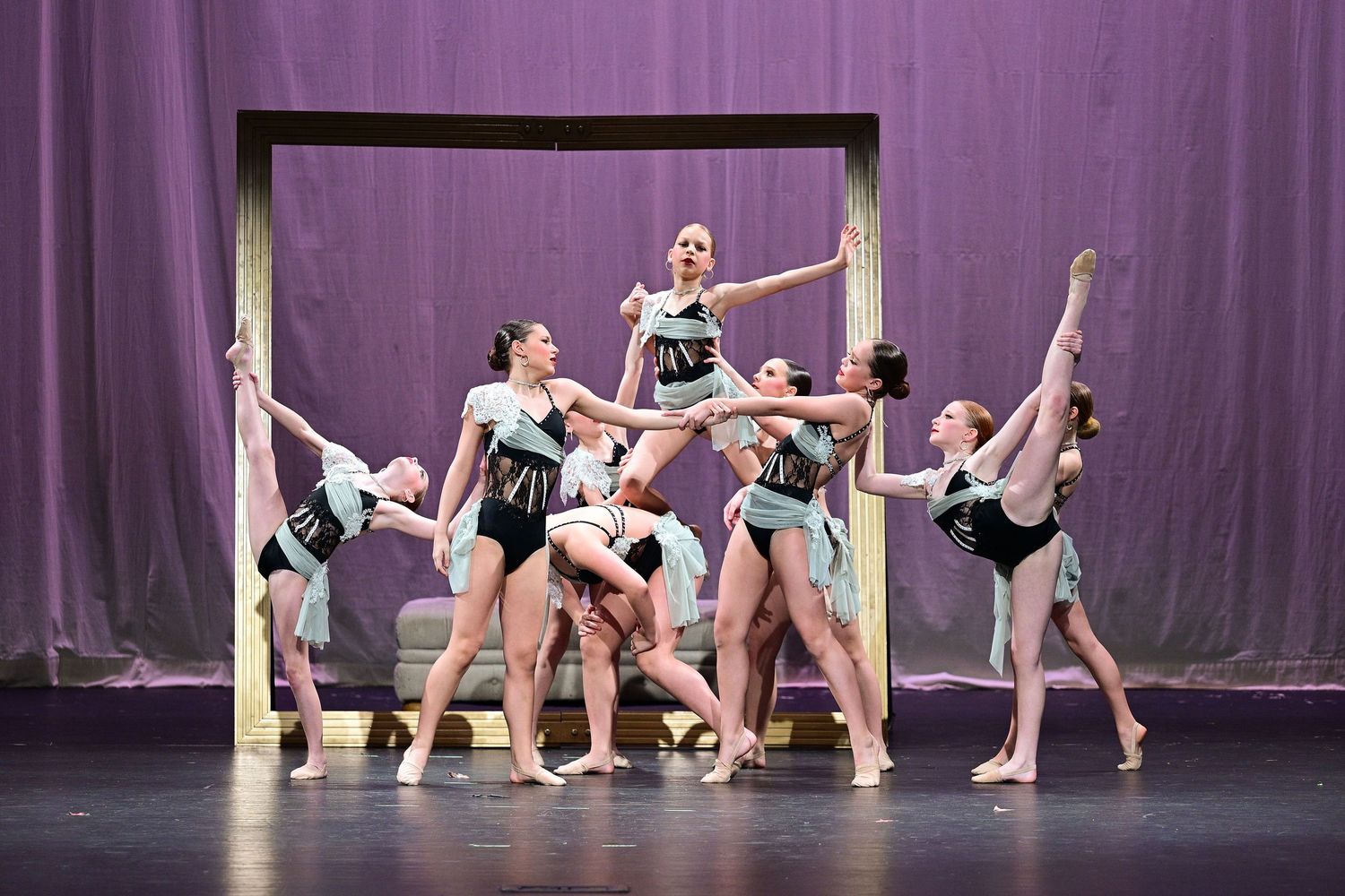 Group of ballet dancers performing a dramatic ensemble pose on stage with purple curtain backdrop.