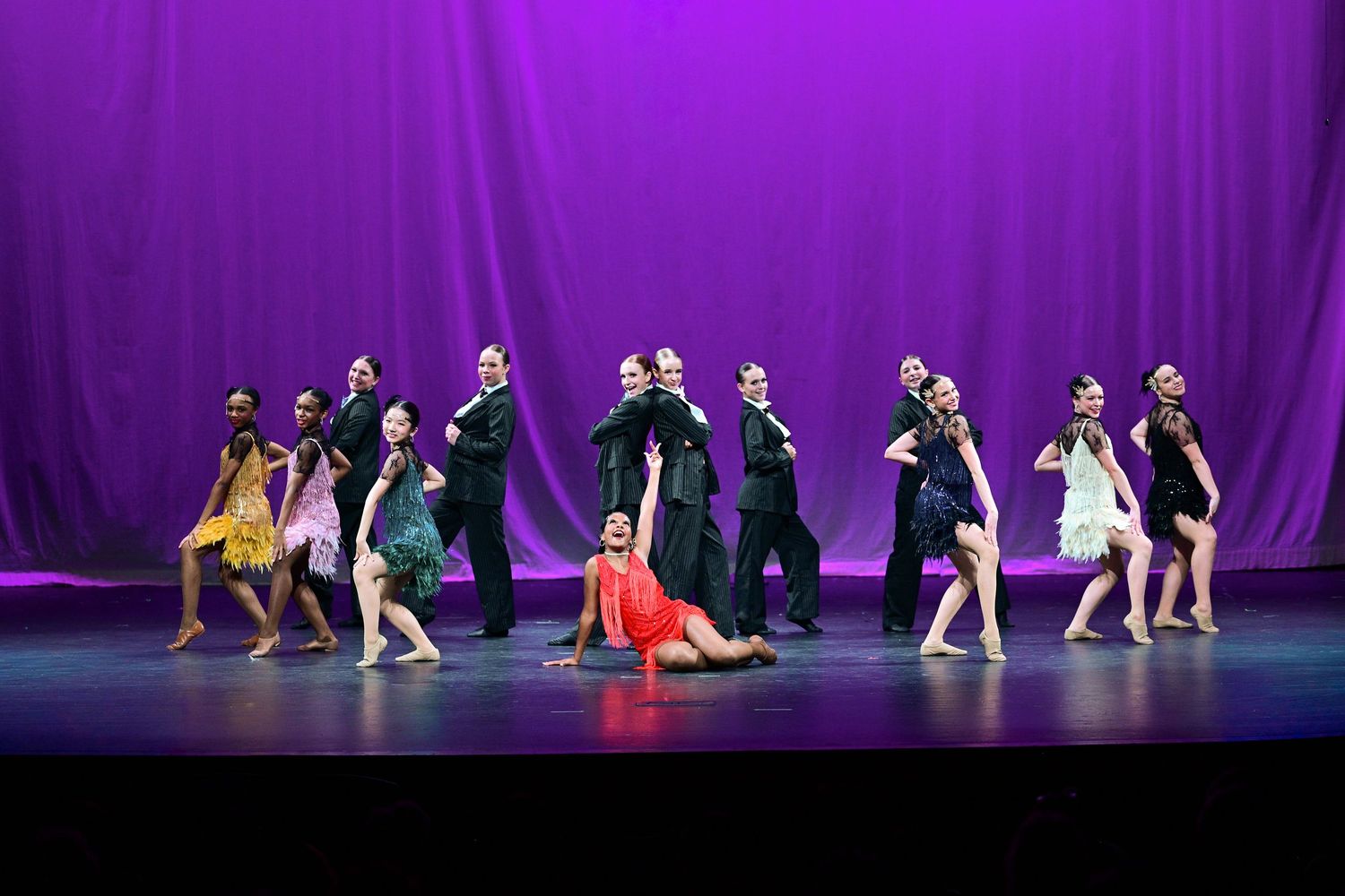 Dancers perform a synchronized routine on stage with dramatic purple lighting and black curtains in the background.