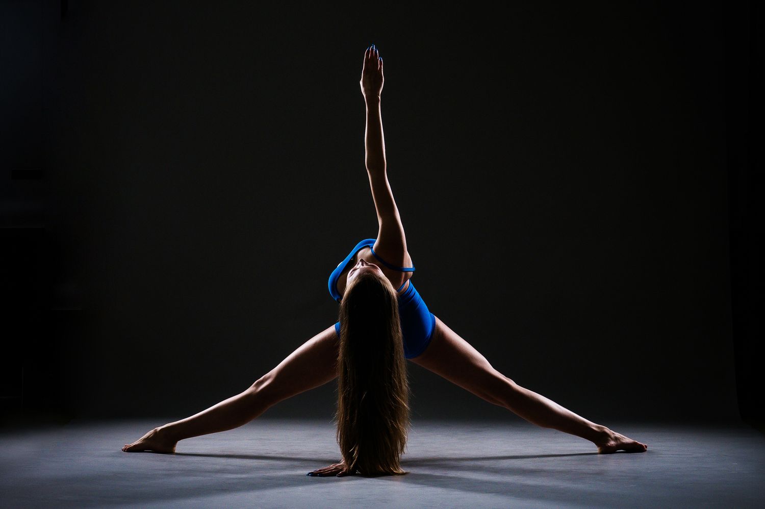 Gymnast in blue leotard performing a split stretch with one arm raised on a dark studio floor.
