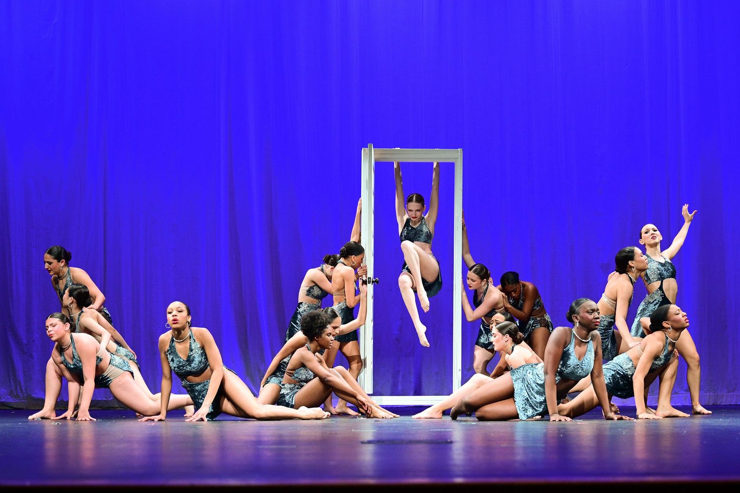 A dance ensemble performs on stage with blue backdrop, featuring an aerial acrobat in a suspended frame.