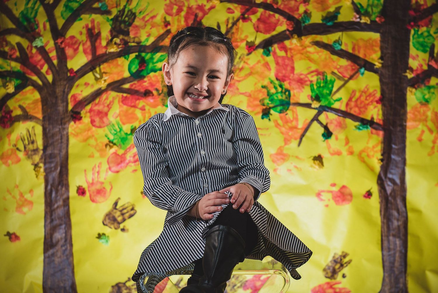 Child in striped sweater laughs while surrounded by autumn-themed backdrop with trees.