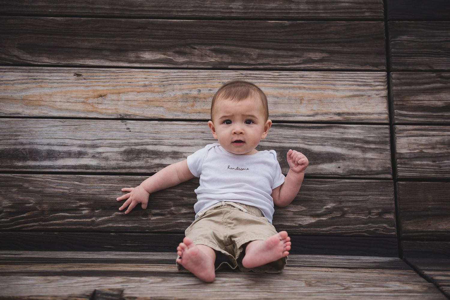 Baby in white onesie sitting against weathered wooden background.