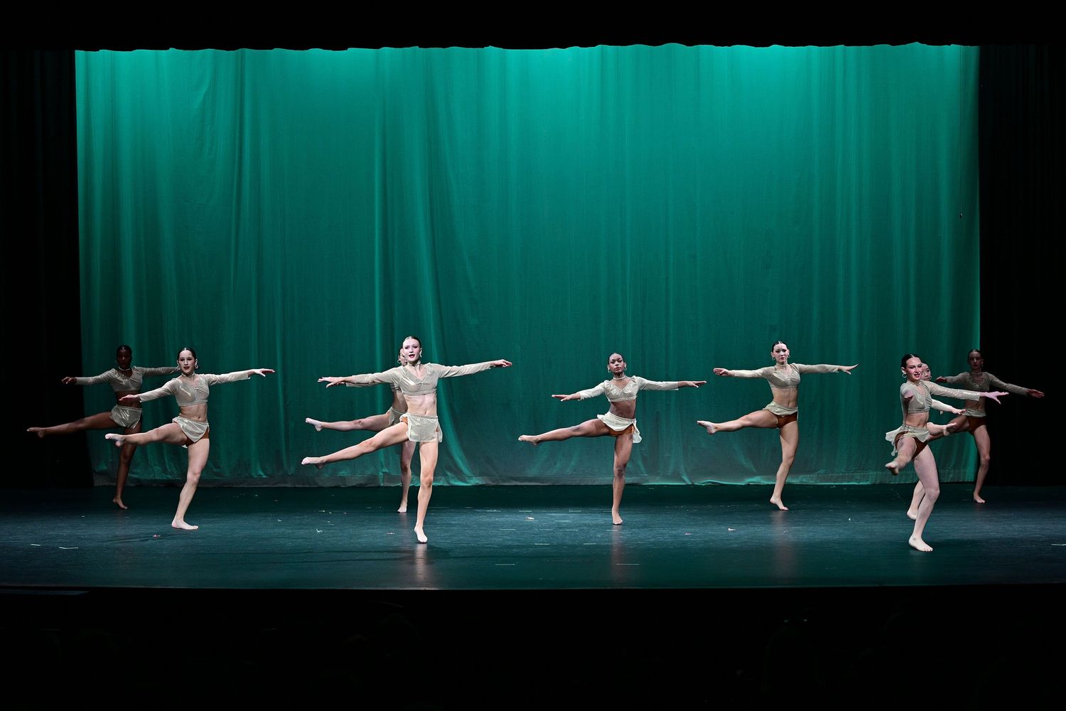 Group of dancers performing synchronized ballet routine on stage with green curtain backdrop.