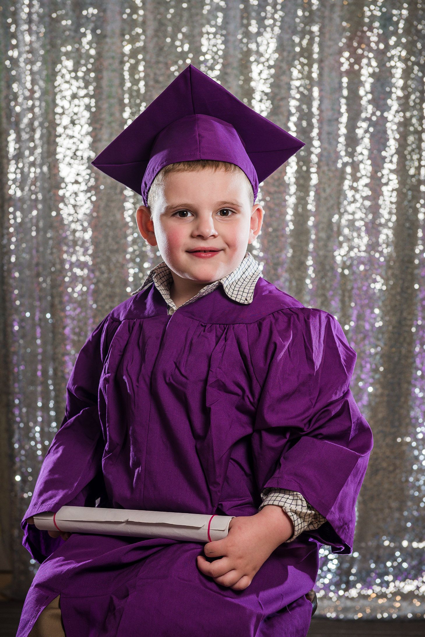 Graduate in purple cap and gown posing against silver sequin backdrop.
