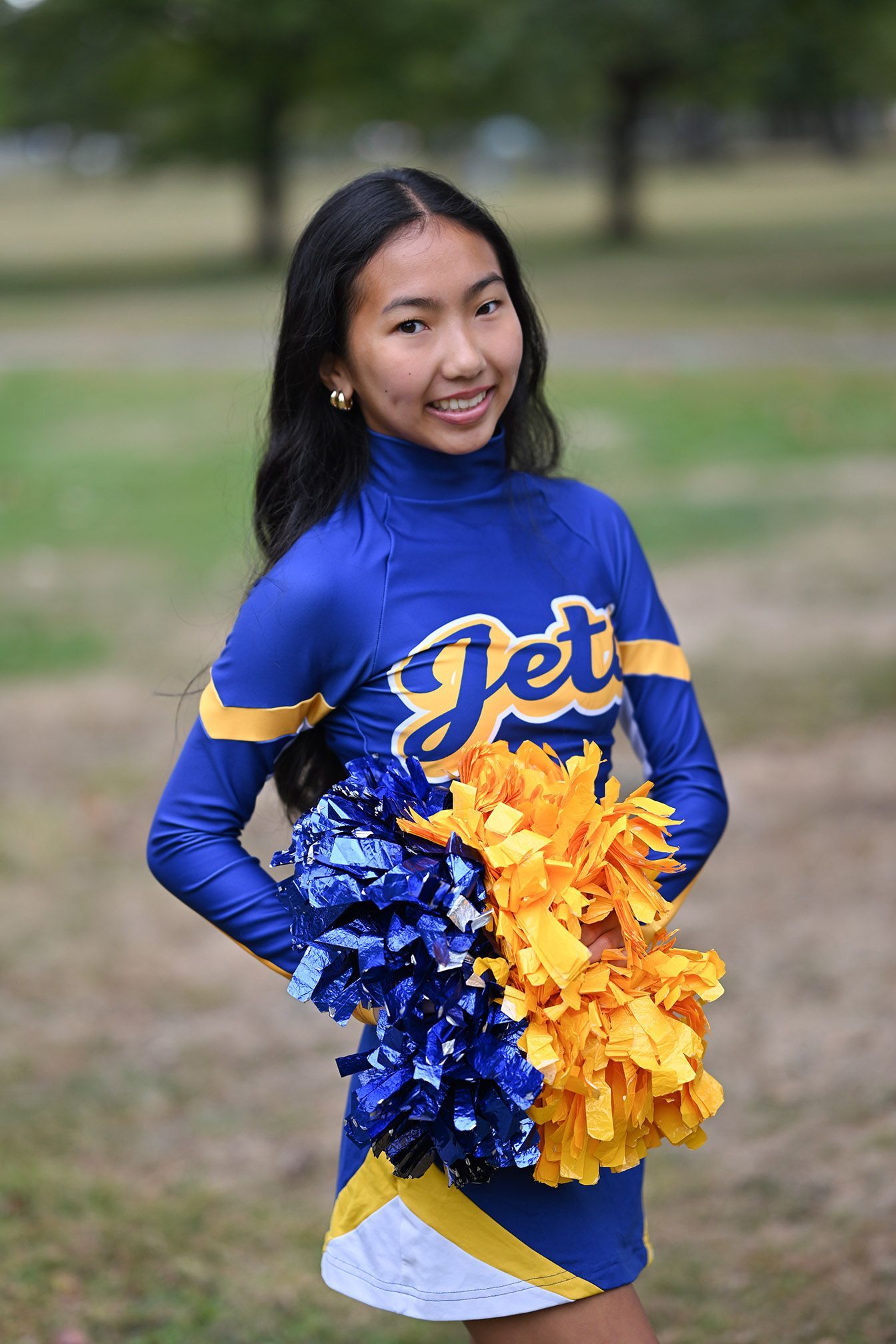 Cheerleader in blue uniform holds blue and yellow pom poms while posing outdoors.