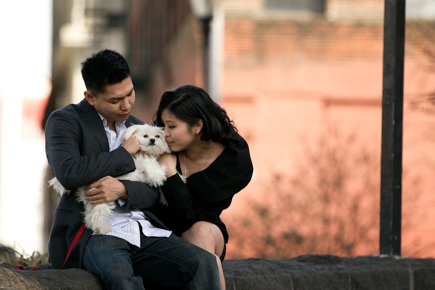 Couple cuddles with small white dog while sitting together outdoors.