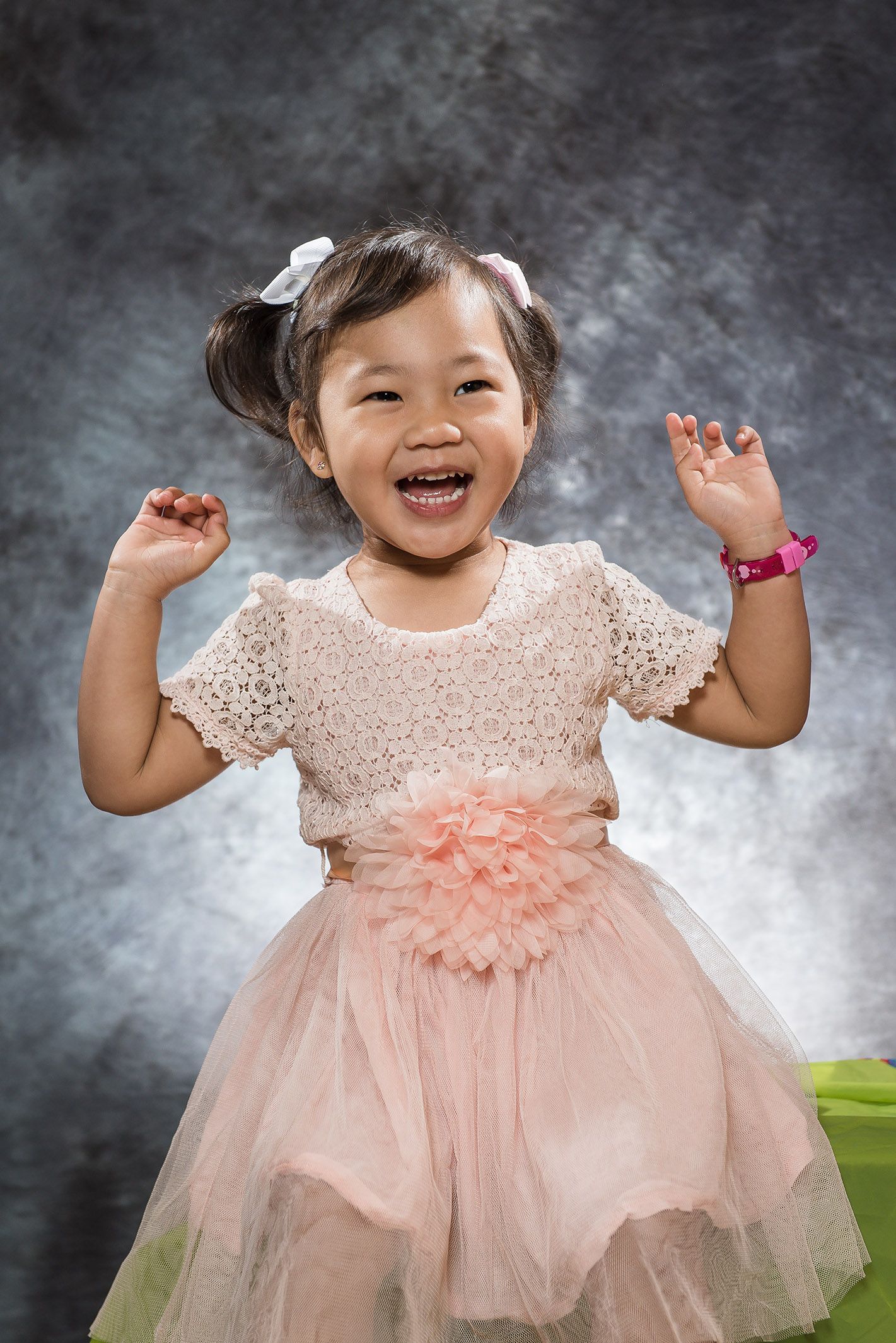 A joyful toddler in a pink tulle dress with lace details dances against a gray backdrop.