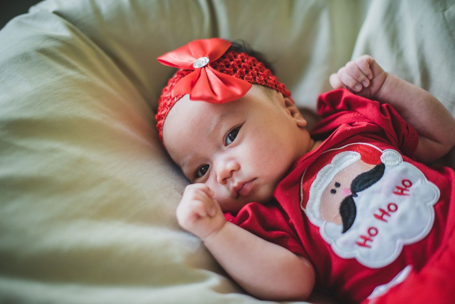 Baby wearing red Santa outfit with matching bow headband lying on cushion.