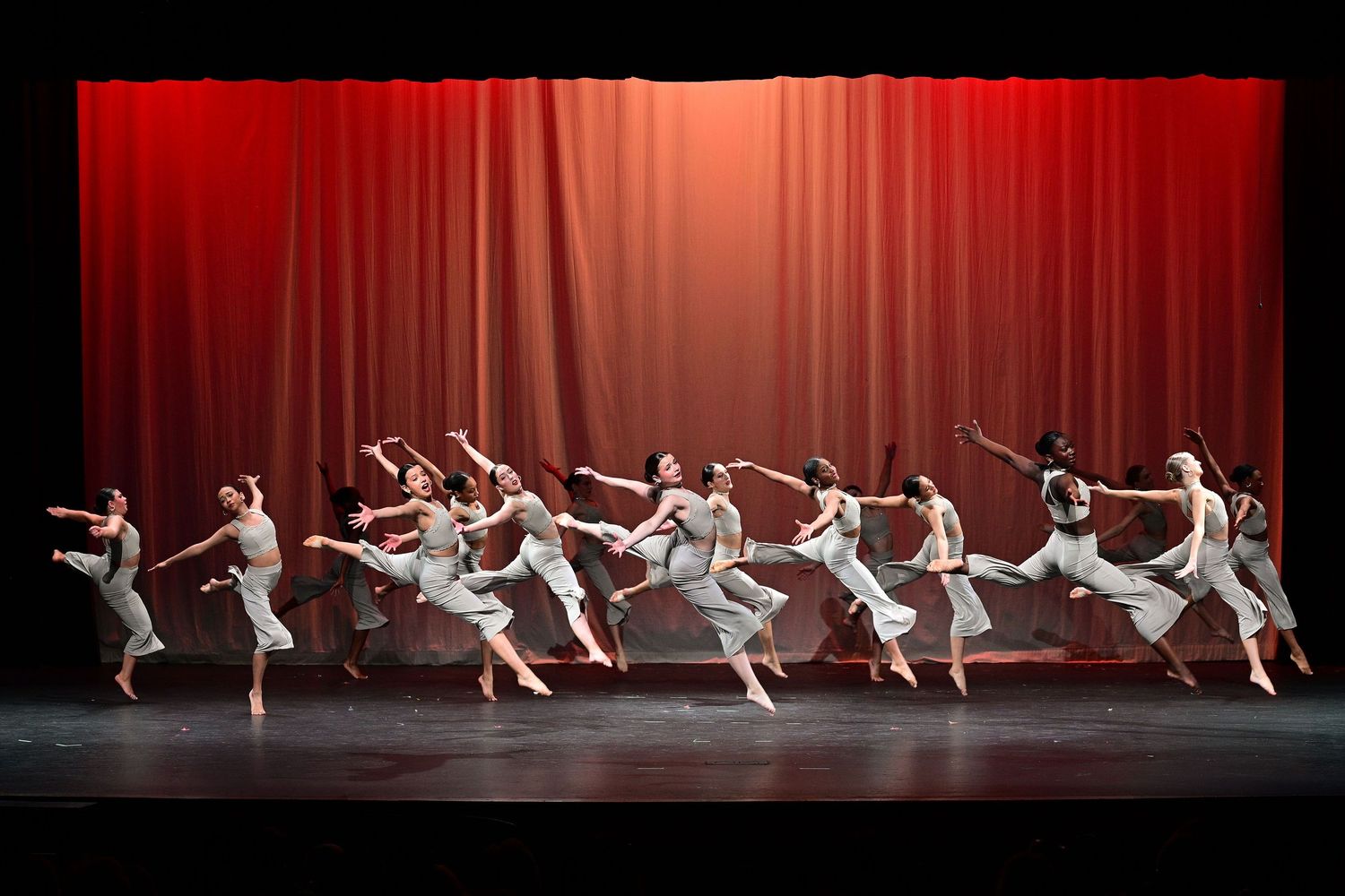 Dancers in white costumes perform a synchronized contemporary ballet routine on a stage with red curtain backdrop.