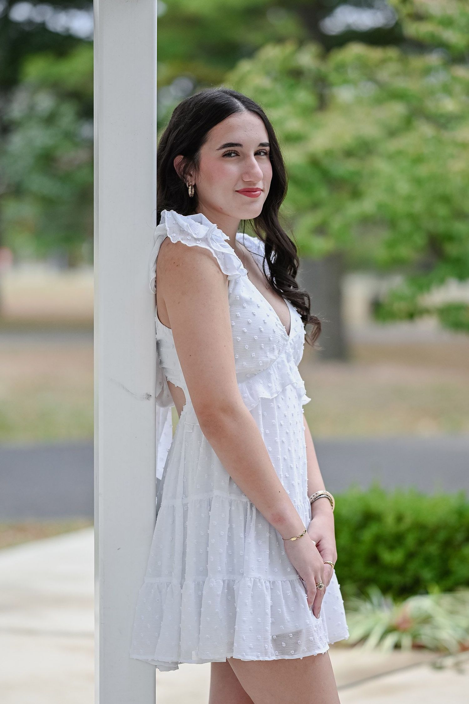 Woman in white ruffled summer dress poses outdoors against green foliage background.
