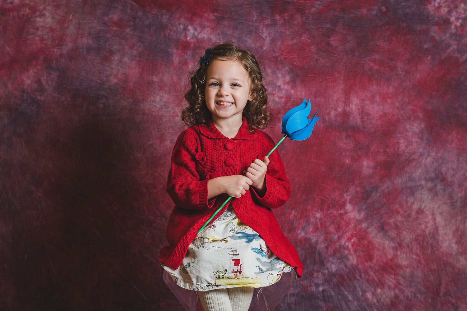 Child in graduation attire celebrates with cake and decorative props against red backdrop.