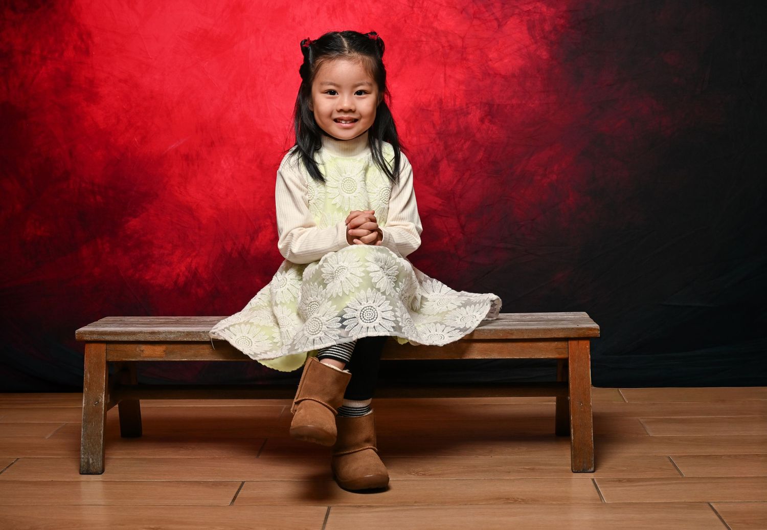 A young child in a white dress sits on a wooden bench against a dramatic red backdrop.
