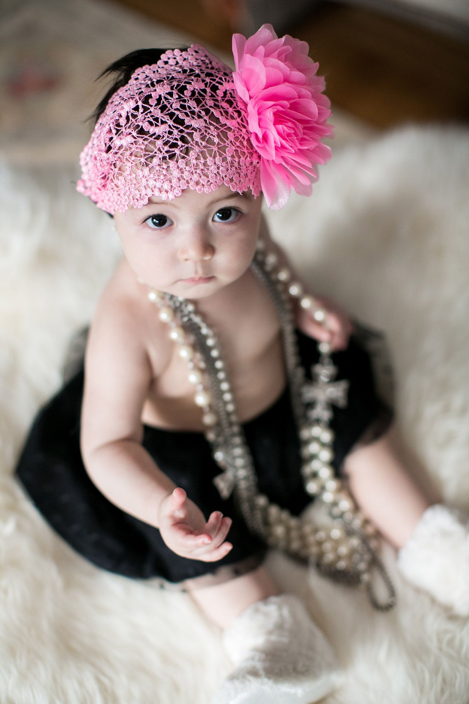 Baby wearing pink headpiece and pearls poses on white fur backdrop.