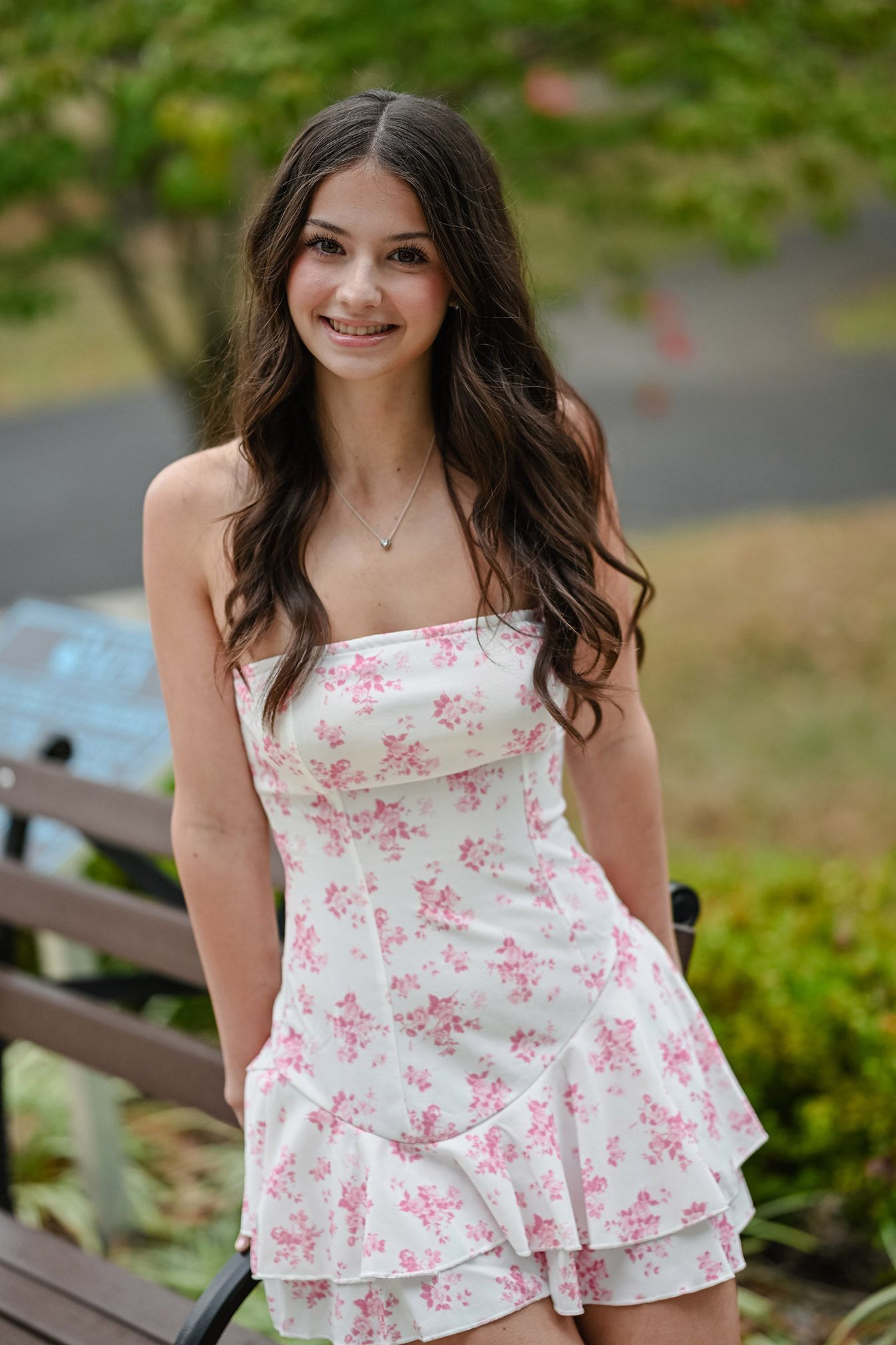 Young woman in floral white and pink top posing on outdoor bench in garden setting.
