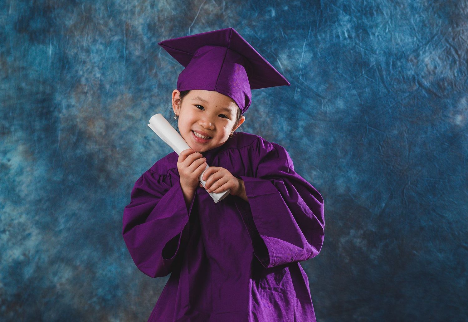 Child wearing purple graduation cap and gown poses playfully against blue backdrop.